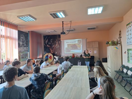 Dzieci siedzą w Sali edukacyjnej przy stołach. Pracownik ZPKWŚ pokazuje na slajdzie bobra i opowiada o nim.
