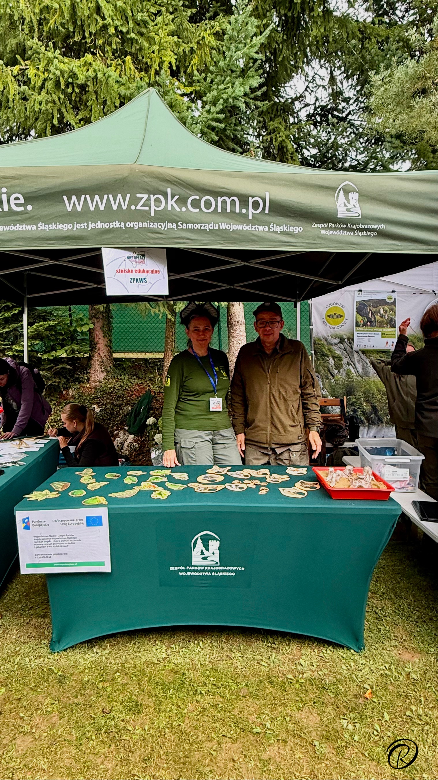 Zdjęcie przedstawia stoisko edukacyjne na świeżym powietrzu, należące do Zespołu Parków Krajobrazowych Województwa Śląskiego. Nad stoiskiem rozpięty jest duży, zielony namiot imprezowy. Pod namiotem, na trawie, stoi długa ława przykryta dopasowanym, zielonym obrusem z logo Zespół Parków Krajobrazowych Województwa Śląskiego. Na stole znajduje się wiele drewnianych plakietek edukacyjnych o różnych kształtach, przedstawiających liście drzew, owoce, a także zwierzęta. Po lewej stronie stołu przypięta jest  pionowa tablica informacyjna z tekstem o projekcie. Za stołem stoją dwie osoby dorosłe: kobieta w zielonej bluzie mundurowej z identyfikatorem na smyczy i mężczyzna w oliwkowym polarze i czapce. Oboje patrzą w stronę aparatu. a mężczyzna uśmiecha się. Za namiotem widać gęste korony drzew iglastych.