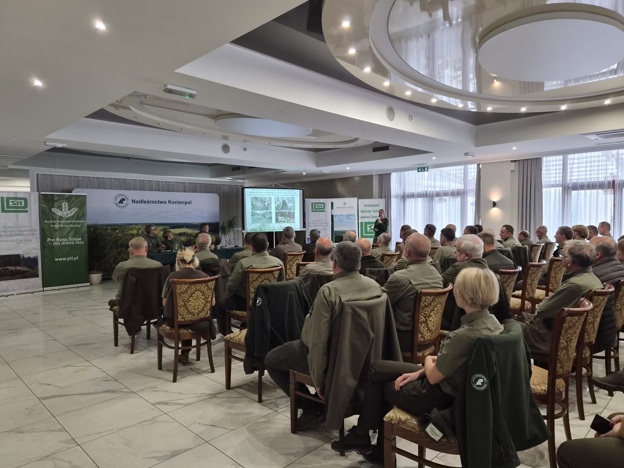Zdjęcie sali konferencyjnej pełnej ludzi siedzących na krzesłach, zwróconych w kierunku prelegentki i trzech osób prowadzących wydarzenie. Sala jest jasna w barwach białych i zielonych.
