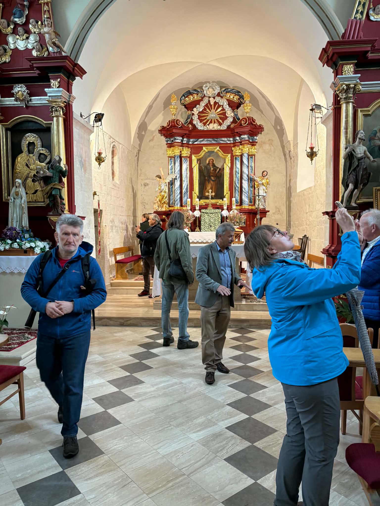 Zdjęcie przedstawia grupę turystów zwiedzających wnętrze jasnego, zabytkowego kościoła. Na pierwszym planie po prawej stronie stoi kobieta w niebieskiej kurtce i szarych spodniach, która z odchyloną do tyłu głową robi zdjęcie telefonem komórkowym w kierunku bogato zdobionego, drewnianego ołtarza. Po lewej stronie pierwszego planu mężczyzna w niebieskiej bluzie z plecakiem idzie w stronę obiektywu. W głębi nawy, na posadzce we wzór szachownicy, widać kilku innych zwiedzających rozmawiających ze sobą oraz przyglądających się wnętrzu. W tle znajduje się ołtarz główny w odcieniach czerwieni, złota i bieli z obrazem religijnym w centralnym punkcie, a po bokach widoczne są dodatkowe zdobienia, rzeźby postaci świętych oraz boczne ołtarze.