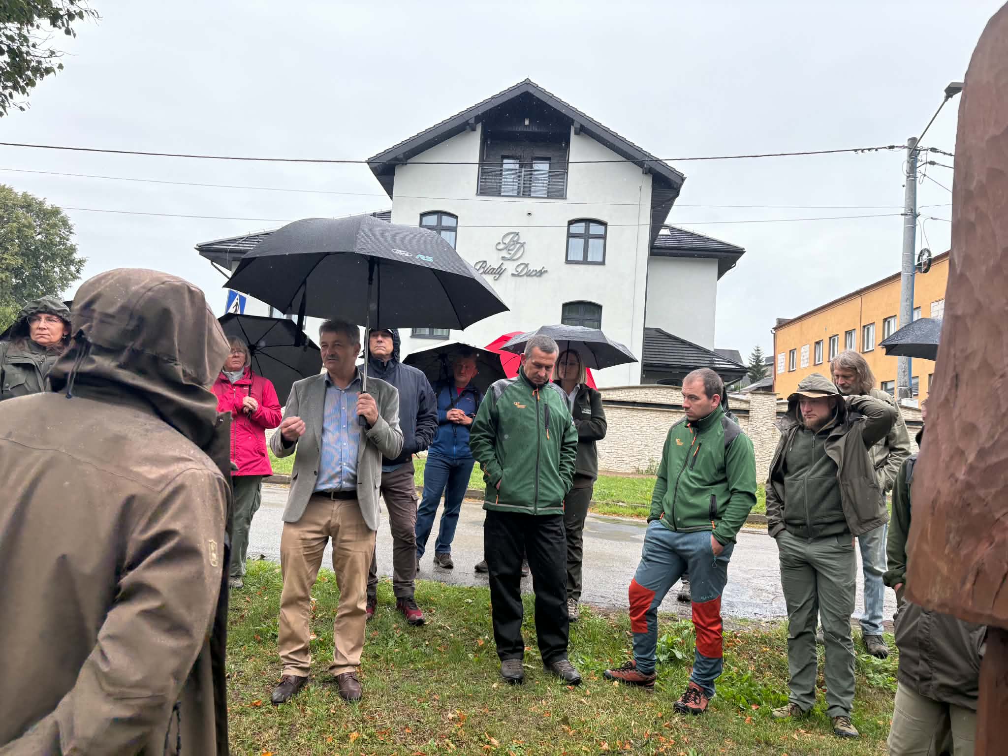 Zdjęcie przedstawia grupę ludzi zgromadzonych na zewnątrz w deszczowy dzień. W centrum uwagi znajduje się mężczyzna w szarej marynarce i beżowych spodniach, trzymający czarny parasol i przemawiający do reszty grupy. Część obecnych osób również osłania się czarnymi parasolami, a niektórzy mają na sobie kurtki z kapturem lub płaszcze przeciwdeszczowe. Mężczyźni stojący bliżej pierwszego planu ubrani są w zielone, sportowo-turystyczne kurtki oraz spodnie do chodzenia po górach. W tle widoczny jest okazały, biały, piętrowy budynek z czarnym dachem i napisem „Biały Dwór” na elewacji, a także mokra jezdnia i pochmurne niebo.