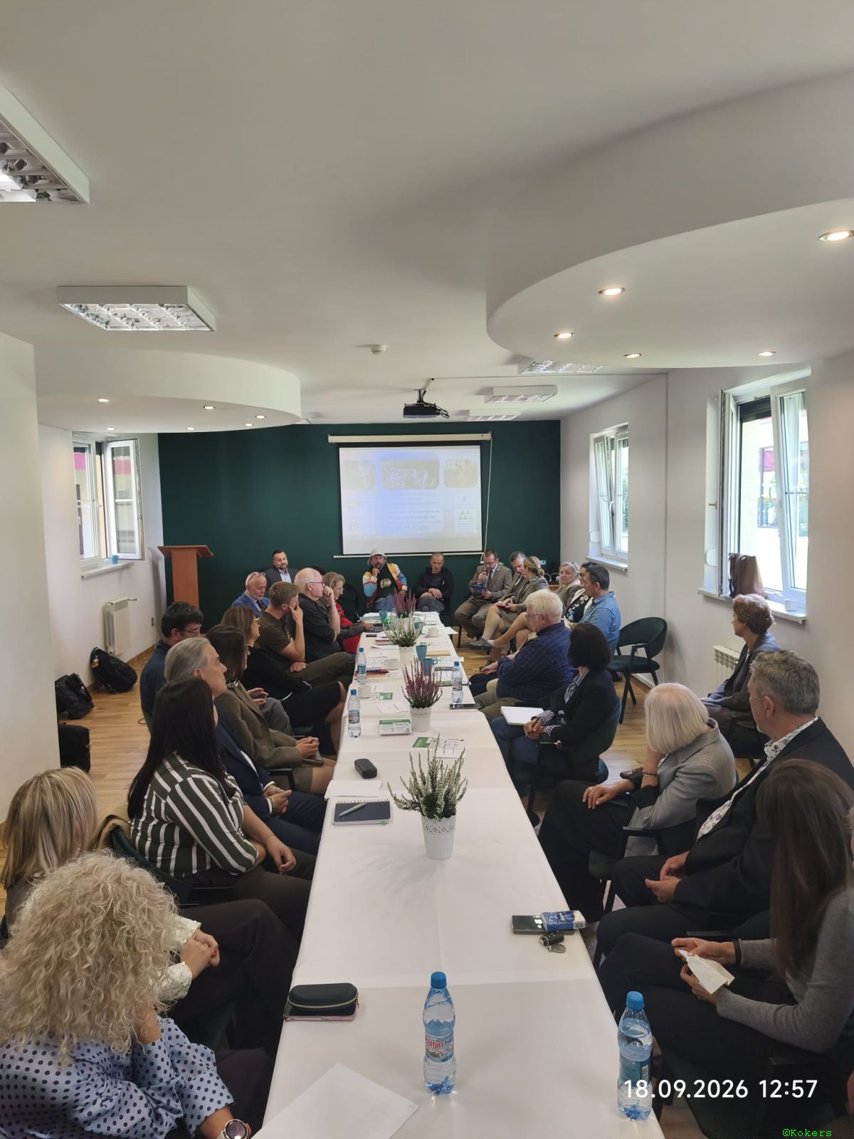 Ujęcie z końca długiego stołu konferencyjnego na około 25 osób siedzących i słuchających prezentacji wyświetlanej z projektora na ścianie w jasnej sali konferencyjnej. Na dole zdjęcia widoczna data: 18.09.2026 12:57.
