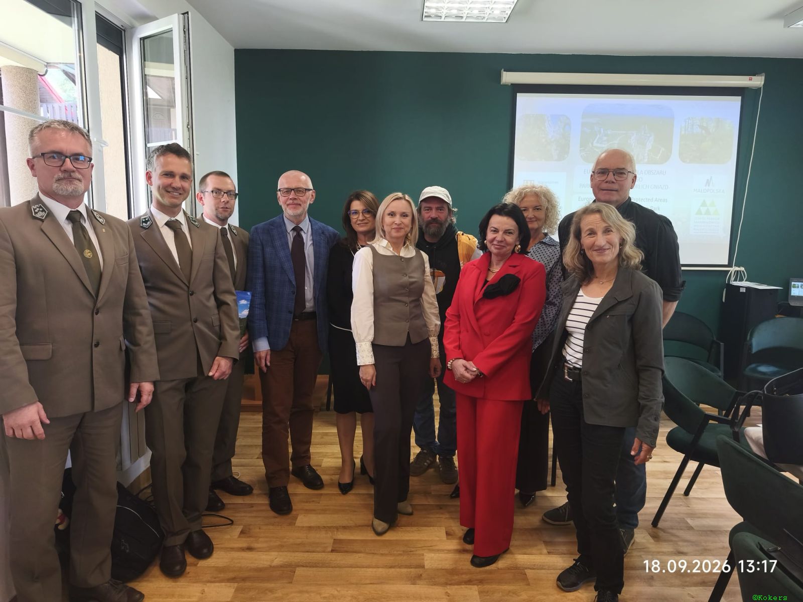 Pamiątkowe zdjęcie grupowe jedenastu osób stojących w sali konferencyjnej przed ekranem projektora. W grupie są strażnicy w oliwkowych mundurach, kobieta w czerwonym garniturze i inni eksperci. Na dole widoczna data: 18.09.2026 13:17.