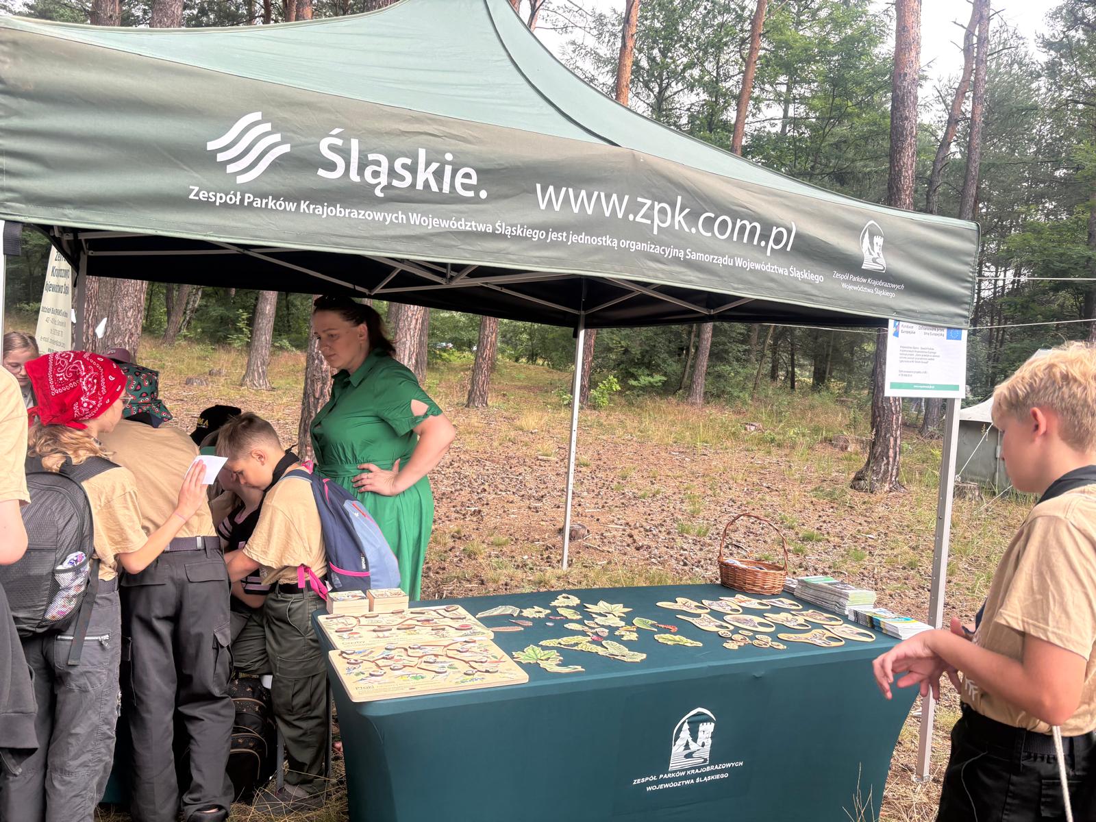 Stoisko edukacyjne Zespołu Parków Krajobrazowych Województwa Śląskiego (ZPKWŚ) w leśnym obozowisku harcerskim. Grupa harcerzy w beżowych mundurach rozmawia z pracownicą ZPKWŚ pod zielonym namiotem promocyjnym, na którego stelażu wisi biały plakat informacyjny Funduszy Europejskich. Na stole nakrytym zielonym obrusem leżą drewniane układanki przyrodnicze, liście oraz broszury edukacyjne