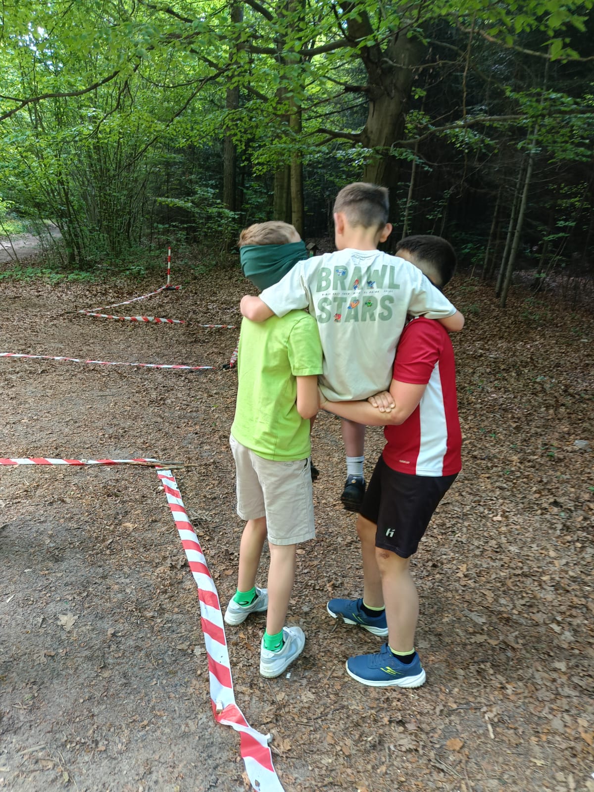 Zdjęcie przedstawia troje dzieci uczestniczących w grze terenowej z przeszkodami na świeżym powietrzu, w lesie. Dwoje dzieci - jedno w jaskrawozielonej koszulce, drugie w czerwono-białej - niesie swojego kolegę w jasnej koszulce na tzw. "krzesełku" splecionym z rąk. Chłopiec po lewej stronie, niosący kolegę, ma przewiązaną na oczach zieloną opaskę. Dzieci stoja na leśnej ścieżce usłanej liśćmi.