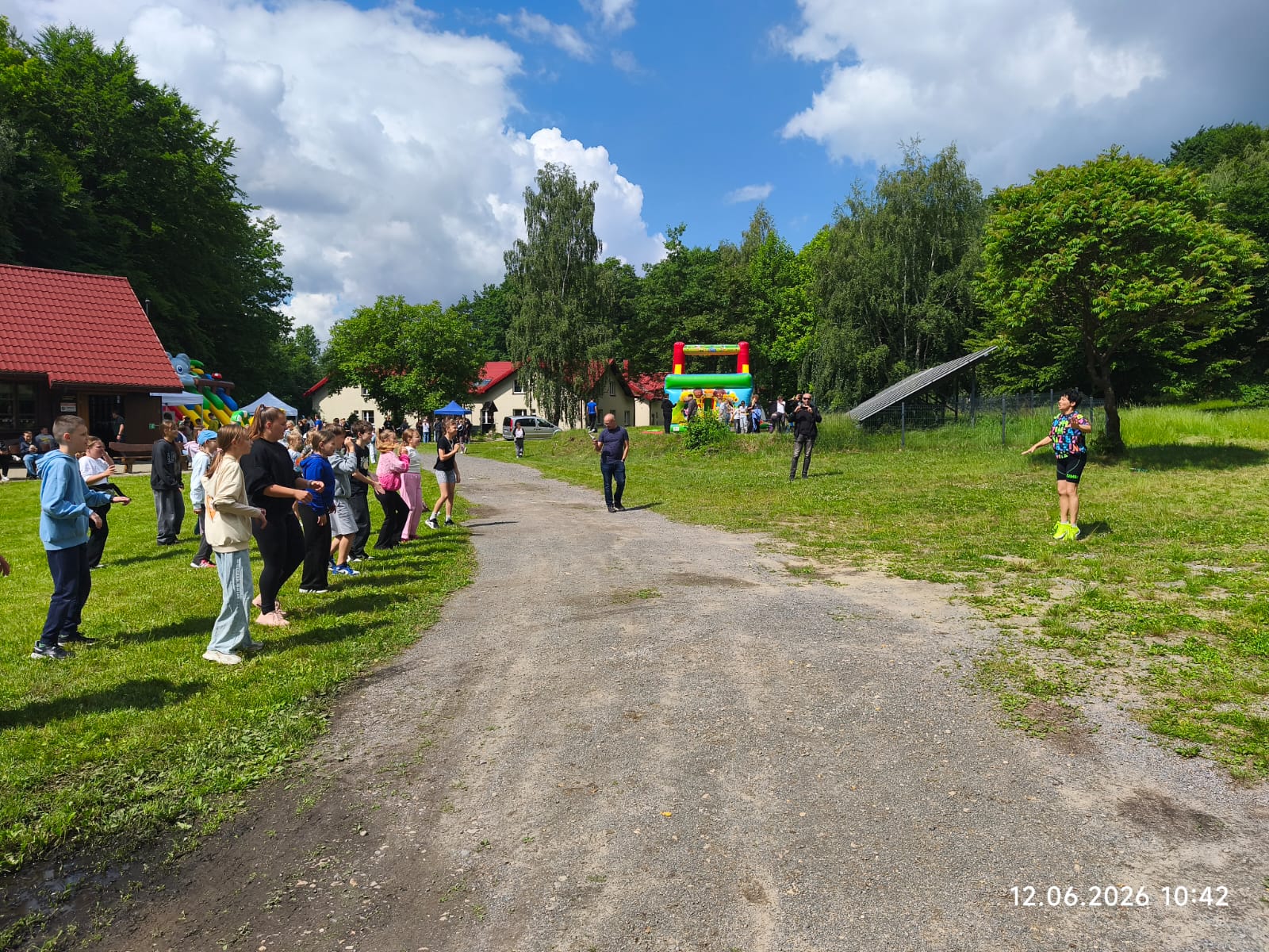 Uczestnicy plenerowego pikniku biorą udział w zorganizowanych zajęciach ruchowych lub tańcu na polanie oddziału ZPKWŚ w Smoleniu. Grupa dzieci i młodzieży stoi na trawniku po lewej stronie, naśladując ruchy animatorki stojącej naprzeciwko nich po prawej stronie. Przez środek polany przebiega szutrowa ścieżka, na której widać przechodniów. W tle widoczne są zabudowania z czerwonymi dachami, kolorowe dmuchane zamki do skakania, panele fotowoltaiczne oraz otaczający teren gęsty las pod częściowo zachmurzonym niebem.
