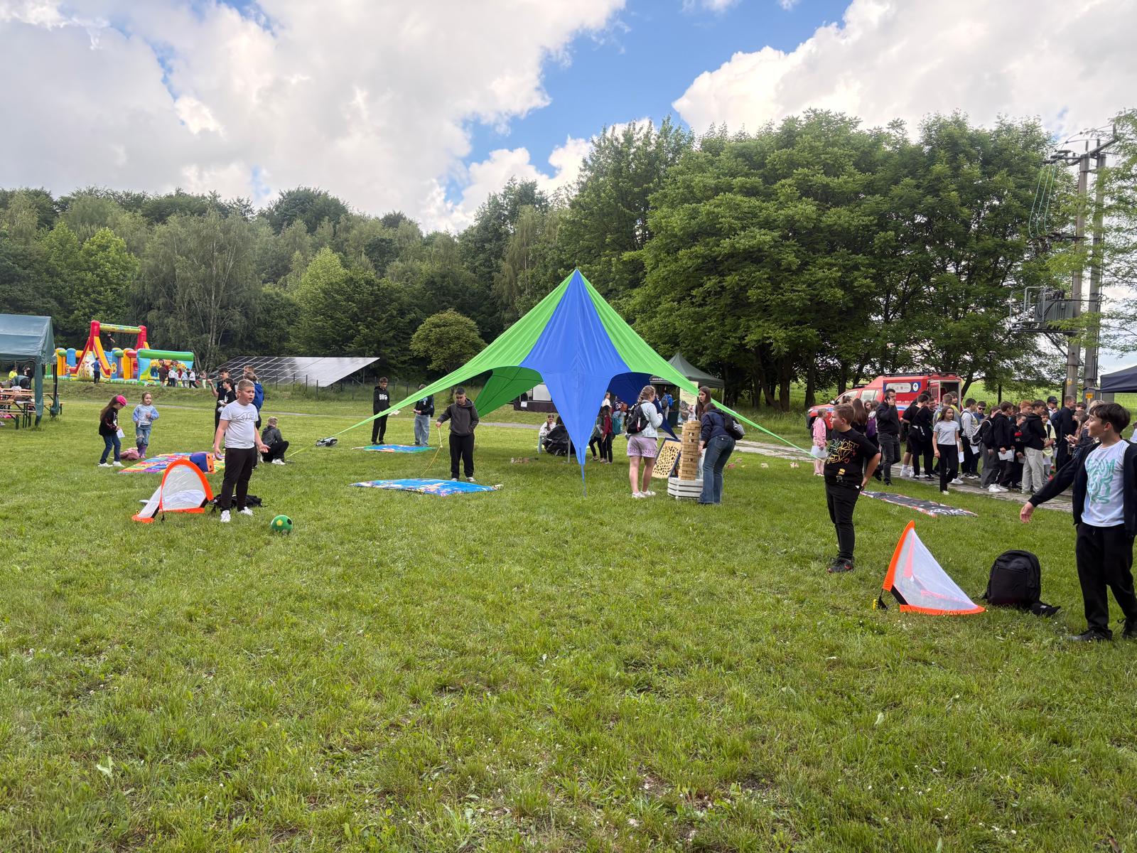 Plenerowy piknik edukacyjny na łące przy oddziale ZPKWŚ w Smoleniu. W centrum stoi charakterystyczny niebiesko-zielony namiot w kształcie gwiazdy, pod którym dzieci układają wieżę z gigantycznych klocków. Na trawniku rozstawiono minibramki do piłki nożnej, a po lewej stronie widać rozłożone maty animacyjne oraz kolorowy dmuchany zamek w tle. Po prawej stronie grupa młodzieży gromadzi się przy czerwonym wozie strażackim. W tle widoczny jest las i zamontowane na gruncie panele fotowoltaiczne.