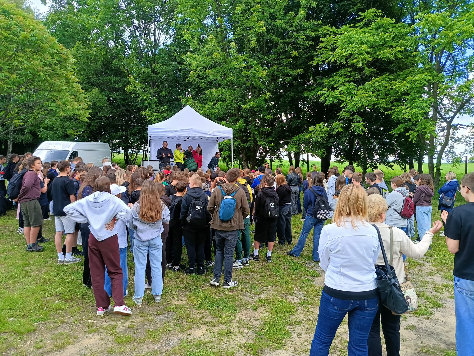 Duża grupa dzieci i młodzieży zgromadziła się na trawiastej polanie przy oddziale ZPKWŚ w Smoleniu, uczestnicząc w plenerowym wydarzeniu. Uczestnicy stoją tyłem do obiektywu i patrzą w stronę białego namiotu ekspozycyjnego, który pełni funkcję sceny. Pod namiotem stoi kilka dorosłych osób prowadzących spotkanie, w tym kobieta w jaskrawożółtej bluzie mówiąca przez mikrofon. Po lewej stronie w tle zaparkowany jest biały samochód dostawczy, a całe wydarzenie otaczają gęste, zielone drzewa.