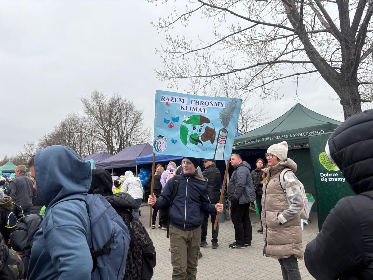 Na zdjęciu przedstawiona jest scena z wydarzenia plenerowego odbywającego się w pochmurny dzień. W centrum kadru stoi młody chłopak ubrany w granatową kurtkę i czapkę, trzymający w obu rękach drewniane kijki, do których przymocowany jest ręcznie wykonany transparent. Transparent ma jasnoniebieskie tło i widnieje na nim napis: „RAZEM CHROŃMY KLIMAT”. Poniżej znajduje się rysunek Ziemi z zaznaczonymi elementami przyrody oraz kominem emitującym dym, co symbolizuje zagrożenia dla środowiska. Obok planety widoczne są kolorowe motyle. Wokół chłopaka znajduje się wiele osób – dorośli i dzieci – ubranych w zimowe kurtki, czapki i plecaki. Po prawej stronie stoi kobieta w jasnej, beżowej kurtce i czapce, obserwująca wydarzenie. Po lewej stronie widać osoby odwrócone tyłem, również z plecakami. W tle ustawione są namioty wystawowe w różnych kolorach, m.in. zielony namiot z napisem „Kasa Rolniczego Ubezpieczenia Społecznego”. Pod namiotami gromadzą się uczestnicy wydarzenia, rozmawiają i oglądają przygotowane stoiska.