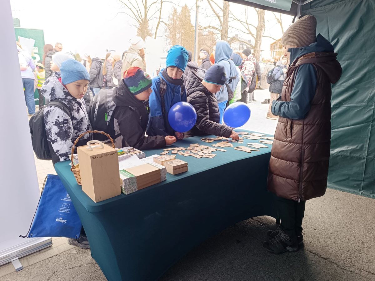 Zdjęcie przedstawia scenę na świeżym powietrzu podczas wydarzenia plenerowego, jest  chłodny dzień (uczestnicy są ubrani w zimowe kurtki, czapki i szaliki). Na pierwszym planie znajduje się stoisko przykryte zielonym obrusem, ustawione pod namiotem. Przy stole stoi grupa kilku dzieci w wieku szkolnym. Mają na sobie zimowe ubrania: puchowe kurtki, czapki i plecaki. Troje z nich trzyma niebieskie balony. Dzieci pochylają się nad stołem i oglądają lub układają drewniane elementy – małe, płaskie klocki i większe wycięte kształty, przypominające puzzle lub edukacyjną układankę. Po prawej stronie stołu stoi dorosła osoba (prawdopodobnie prowadząca stoisko), ubrana w długą, brązową kamizelkę, zieloną kurtkę i czapkę. Osoba ta obserwuje dzieci i jest zwrócona w ich stronę. Na stole, oprócz drewnianych elementów, znajdują się również inne przedmioty: małe pudełka, stosy ulotek lub kart oraz koszyk. Z przodu stołu stoi większe drewniane pudełko z uchwytem.