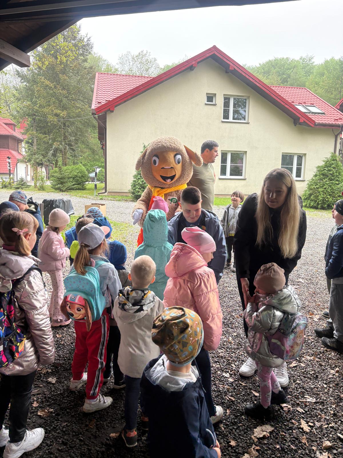 Zdjęcie przedstawia grupę dzieci i dorosłych zgromadzonych na świeżym powietrzu, przed budynkiem mieszkalnym z jasną elewacją i czerwonym dachem. Główną atrakcją spotkania jest osoba przebrana w duży, pluszowy kostium brązowej owieczki (maskotkę), wokół której gromadzą się zaciekawione dzieci ubrane w kolorowe kurtki, czapki i plecaki. Obok dzieci stoi opiekunka, która rozmawia z jednym z maluchów, a w tle widać innych dorosłych obserwujących sytuację. Całość ma charakter zorganizowanej wycieczki, pikniku.